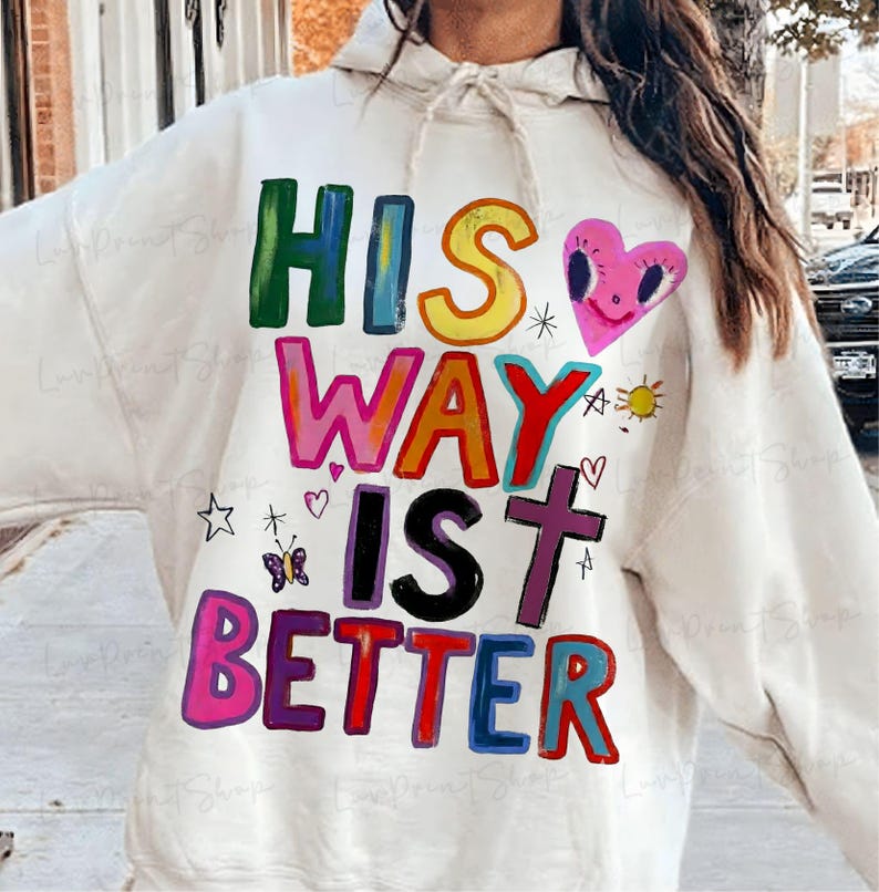 May include: Cream-colored hoodie with the text "HIS WAY IS BETTER" in colorful, block letters. The design includes a pink heart, a cross, a sun, and other small illustrations. The hoodie has a drawstring hood.