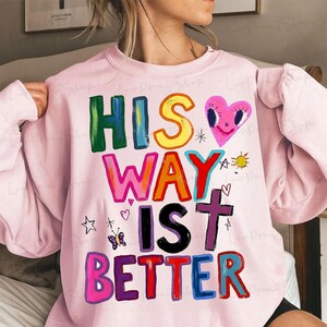 May include: A light pink sweatshirt with the text "HIS WAY IS BETTER" in colorful, block letters. The design includes a pink heart with a smiling face, a cross, a butterfly, and other small graphics.