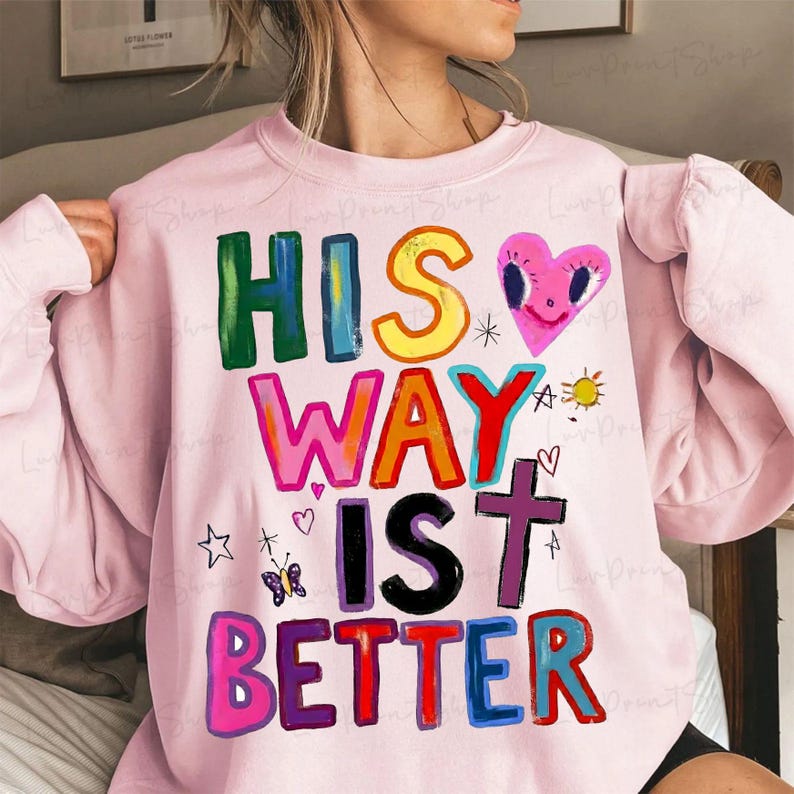 May include: A light pink sweatshirt with the text "HIS WAY IS BETTER" in colorful, block letters. The design includes a pink heart with a smiling face, a cross, a butterfly, and other small graphics.
