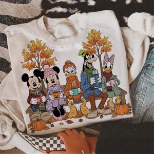 May include: Cream-colored sweatshirt featuring a fall scene with cartoon characters Mickey Mouse, Minnie Mouse, Donald Duck, Goofy, and Daisy Duck. They are sitting on a bench, holding mugs, with pumpkins and autumn leaves.