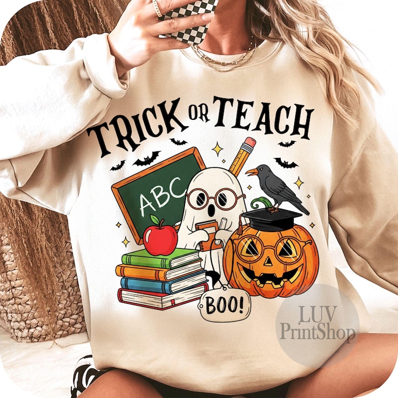 May include: Cream-colored sweatshirt with a Halloween-themed graphic. The design includes a ghost, pumpkin, books, and the words "Trick or Teach." A pencil, apple, and crow are also featured in the illustration. Bats and stars complete the design.