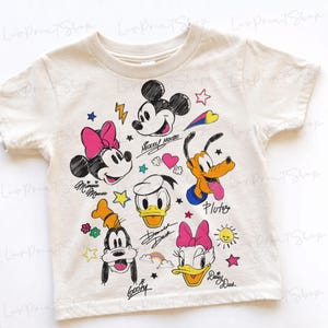 Puede incluir: Camiseta de color crema con ilustraciones de dibujos animados de Mickey Mouse, Minnie Mouse, Donald Duck, Daisy Duck, Goofy y Pluto. El dise&ntilde;o incluye acentos coloridos como lazos rosas, estrellas amarillas y un arco&iacute;ris. Los nombres de los personajes est&aacute;n escritos en cursiva.