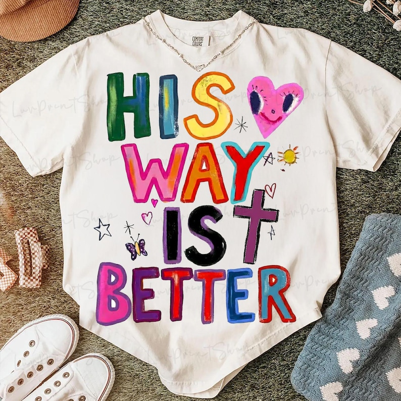 May include: Off-white t-shirt with the phrase "HIS WAY IS BETTER" in colorful, block letters. The design includes a pink heart, a cross, a butterfly, and other small illustrations. The shirt has short sleeves and a crew neck.