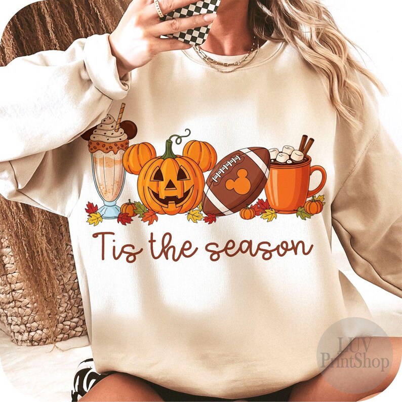 May include: A cream-coloured sweatshirt featuring a seasonal graphic. The design includes a milkshake, a carved pumpkin, a football, and a mug of hot chocolate. The text "Tis the season" is written in brown script across the bottom.