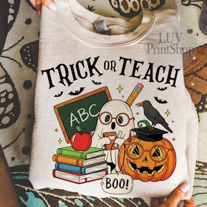 May include: Beige sweatshirt with a Halloween-themed graphic. The design features the text "TRICK OR TEACH" above a ghost wearing glasses, a chalkboard with "ABC", a pumpkin with glasses, books, an apple, and a crow.