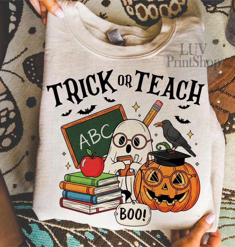 May include: Beige sweatshirt with a Halloween-themed graphic. The design features the text "TRICK OR TEACH" above a ghost wearing glasses, a chalkboard with "ABC", a pumpkin with glasses, books, an apple, and a crow.