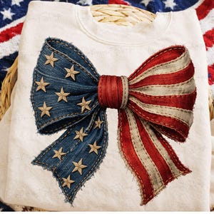 May include: A white sweatshirt featuring a large bow design. The bow is composed of denim-like fabric with gold stars and red and white striped fabric, evoking an American flag theme. The bow is centered on the front of the sweatshirt.