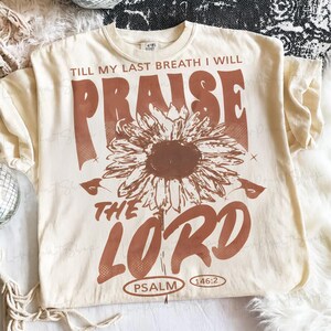 May include: Cream-colored t-shirt with a brown sunflower graphic and text that reads "TILL MY LAST BREATH I WILL PRAISE THE LORD PSALM 146:2". The design is centered on the front of the shirt.