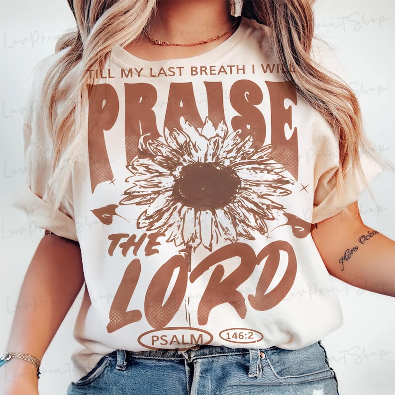 May include: Cream-colored t-shirt with a brown sunflower graphic and the words "PRAISE THE LORD" in a bold, vintage font. The text "TILL MY LAST BREATH I WILL" is above the sunflower, and "PSALM 146:2" is below.