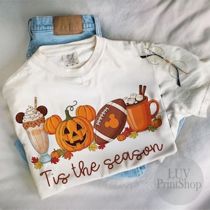 May include: Cream t-shirt with a seasonal graphic. The design includes a milkshake, pumpkins, a football, and a mug of hot chocolate. The phrase "Tis the season" is written in brown script. The shirt is placed on blue jeans.
