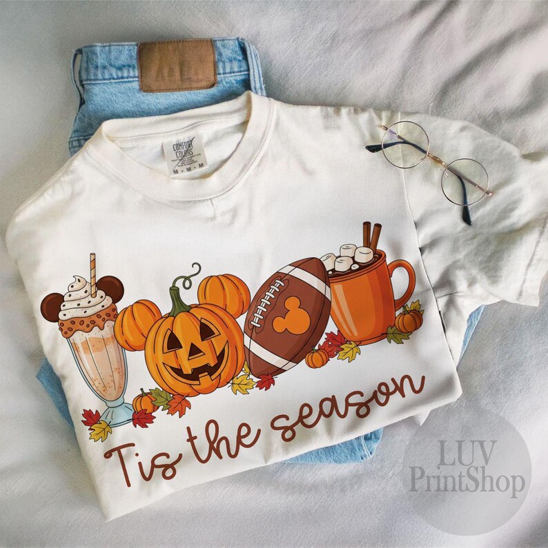 May include: Cream t-shirt with a seasonal graphic. The design includes a milkshake, pumpkins, a football, and a mug of hot chocolate. The phrase "Tis the season" is written in brown script. The shirt is placed on blue jeans.