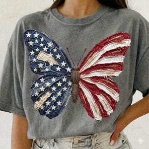 Puede incluir: Camiseta gris con un dise&ntilde;o de mariposa. Las alas de la mariposa est&aacute;n hechas de tela de retazos roja, blanca y azul, imitando la bandera estadounidense. El cuerpo de la mariposa es de color marr&oacute;n texturizado.