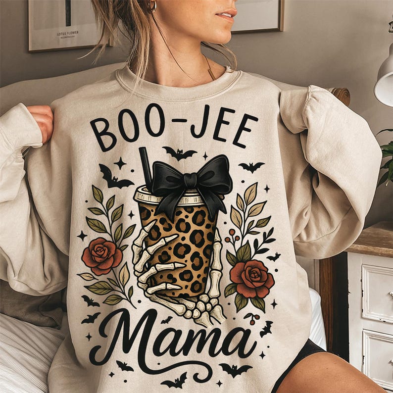 Op de afbeelding: Beige sweatshirt met een Halloween-thema. Het ontwerp toont de tekst "BOO-JEE Mama" boven een beker met luipaardprint die wordt vastgehouden door een skelet, een zwarte strik, rozen, vleermuizen en bladeren.