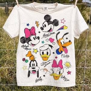 Puede incluir: Camiseta de color crema con ilustraciones de dibujos animados de Mickey Mouse, Minnie Mouse, Donald Duck, Daisy Duck, Goofy y Pluto. La camiseta tiene acentos coloridos y nombres de personajes manuscritos.