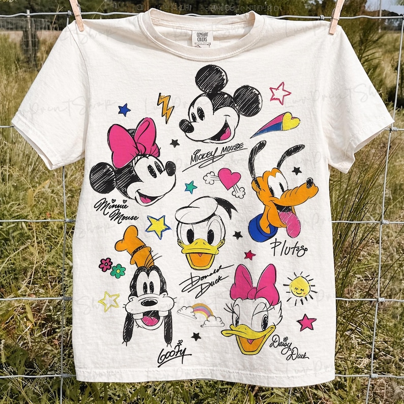 Puede incluir: Camiseta de color crema con ilustraciones de dibujos animados de Mickey Mouse, Minnie Mouse, Donald Duck, Daisy Duck, Goofy y Pluto. La camiseta tiene acentos coloridos y nombres de personajes manuscritos.