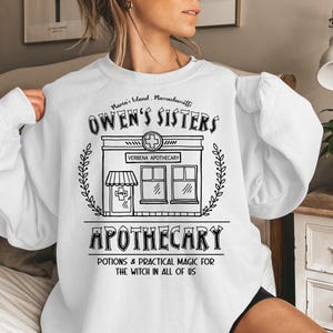 以下が含まれることがあります： 黒い薬局のグラフィックが特徴の白いスウェットシャツ。「OWEN'S SISTERS APOTHECARY」と「POTIONS & PRACTICAL MAGIC FOR THE WITCH IN ALL OF US」の文字。デザインには薬局の十字記号が含まれています。