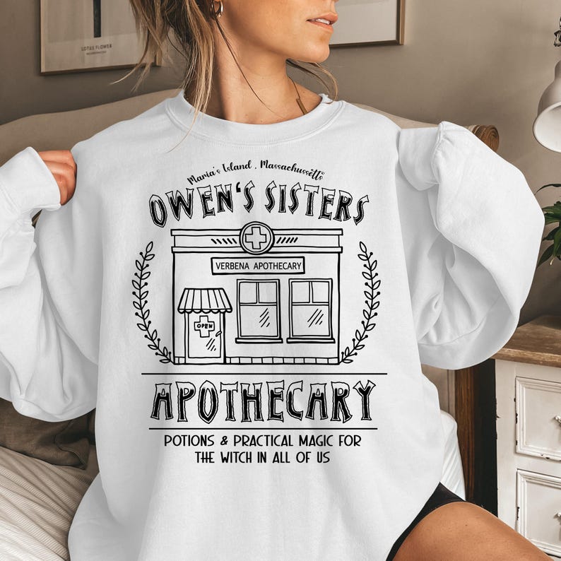 以下が含まれることがあります： 黒い薬局のグラフィックが特徴の白いスウェットシャツ。「OWEN'S SISTERS APOTHECARY」と「POTIONS & PRACTICAL MAGIC FOR THE WITCH IN ALL OF US」の文字。デザインには薬局の十字記号が含まれています。