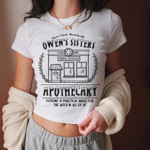 以下が含まれることがあります： 黒いグラフィックデザインの白いクロップトップ。「Owen's Sisters Apothecary」と「Potions & Practical Magic for the Witch in All of Us」のテキストと薬局のイラストが特徴です。