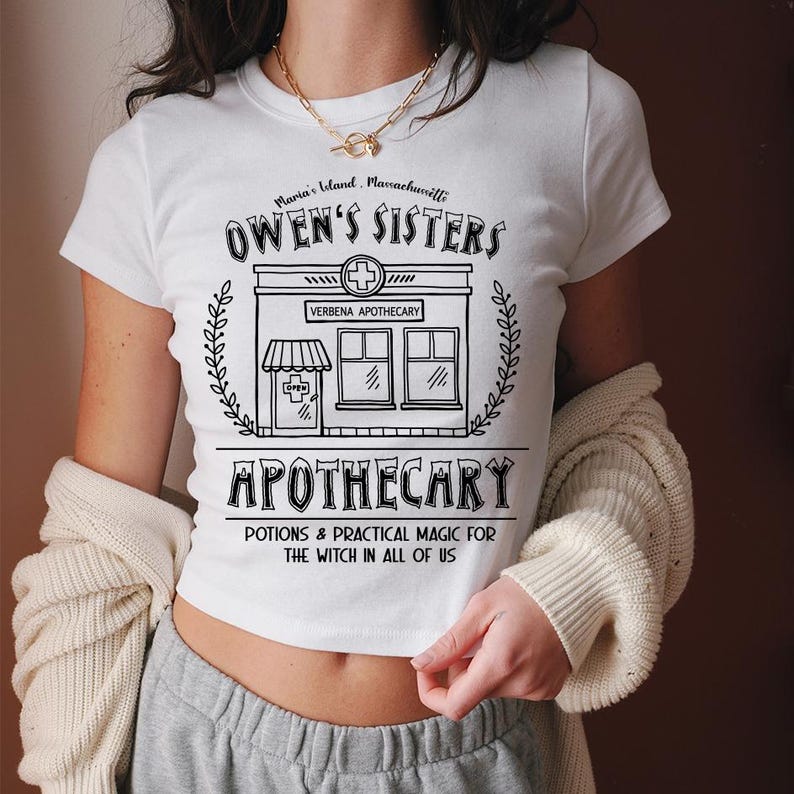 以下が含まれることがあります： 黒いグラフィックデザインの白いクロップトップ。「Owen's Sisters Apothecary」と「Potions & Practical Magic for the Witch in All of Us」のテキストと薬局のイラストが特徴です。