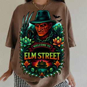 May include: A brown t-shirt featuring a graphic of a character with a fedora and bladed glove, surrounded by colorful floral patterns and a house silhouette. The text reads "WELCOME TO ELM STREET" in bold yellow letters against a dark background.