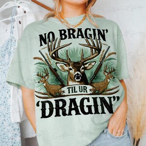 May include: A pale green t-shirt with a deer graphic, antlers, two rifles, and the text "NO BRAGIN' TIL UR DRAGIN'". The design incorporates two smaller deer and a green and brown colour scheme.