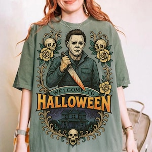 May include: A sage green t-shirt featuring a graphic of Michael Myers holding a knife, surrounded by yellow roses, skulls, and a house illustration. The text reads Welcome to Halloween in bold orange letters, framed by ornate gold scrollwork designs.
