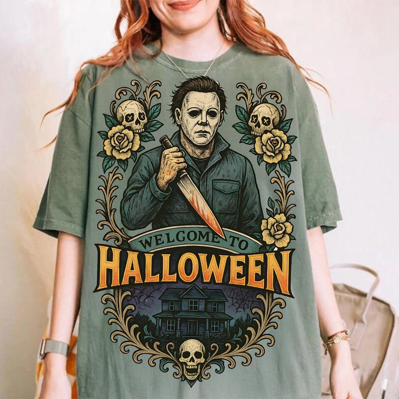 May include: A sage green t-shirt featuring a graphic of Michael Myers holding a knife, surrounded by yellow roses, skulls, and a house illustration. The text reads Welcome to Halloween in bold orange letters, framed by ornate gold scrollwork designs.