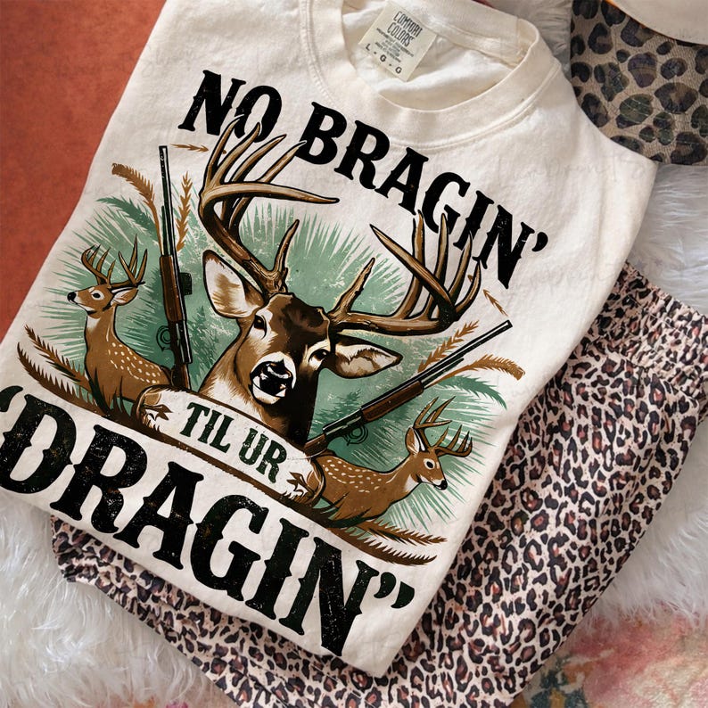 May include: Cream-coloured sweatshirt featuring a deer graphic and the text "NO BRAGIN' TIL UR DRAGIN'". The sweatshirt is paired with leopard print trousers and a matching hat. The design includes deer, rifles, and foliage.