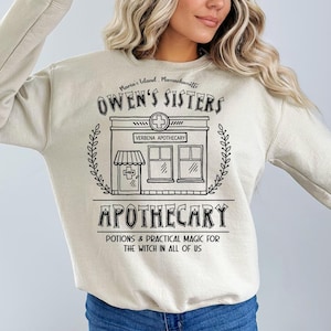 以下が含まれることがあります： クリーム色のスウェットシャツ。黒いグラフィックには「Owen's Sisters Apothecary」と「Maria's Island, Massachusetts」の文字。デザインには店のイラスト、十字のシンボル、そして「Potions & Practical Magic for the Witch in All of Us」というフレーズが含まれています。