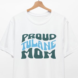 Puede incluir: Camiseta blanca con un gráfico verde y azul que dice "Proud Tulane Mom" en una fuente retro.