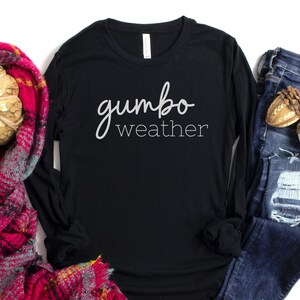 May include: Black long-sleeve shirt with the text "gumbo weather" in white lettering. The shirt is displayed with a red and black plaid scarf, blue jeans, and acorns.
