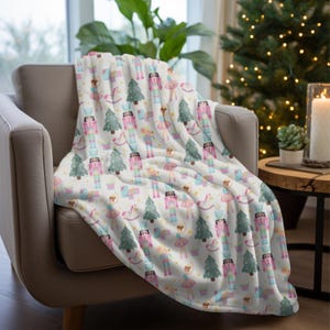 May include: A white throw blanket draped over a chair, featuring a repeating pattern of nutcrackers, ballerinas, Christmas trees, and rocking horses in pastel colors. The blanket is made of a soft material and is suitable for the holiday season.