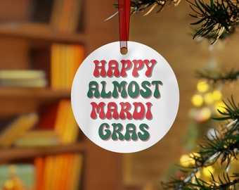 May include: White round Christmas ornament with the text "HAPPY ALMOST MARDI GRAS" in red and green retro font. The ornament hangs from a red ribbon and is displayed on a Christmas tree.