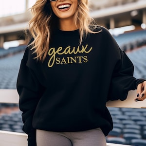 May include: Black sweatshirt with the text "geaux SAINTS" in gold script. The sweatshirt is a crew neck style and appears to be made of a soft material. The image shows a person wearing the sweatshirt.
