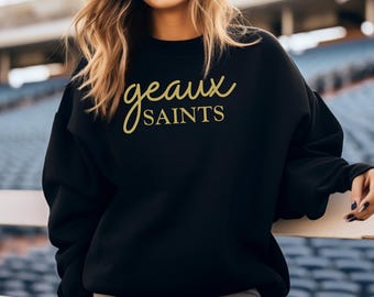 May include: Black sweatshirt with the text "geaux SAINTS" in gold script. The sweatshirt is a crew neck style and appears to be made of a soft material. The image shows a person wearing the sweatshirt.
