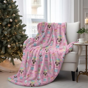 May include: A pink plush throw blanket draped over a white armchair. The blanket features a repeating pattern of cartoon dogs in festive outfits. A Christmas tree with ornaments is in the background, suggesting a holiday theme.