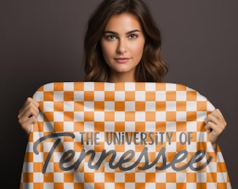 May include: A woman holds up an orange and white checkered blanket with the text "The University of Tennessee" in black lettering.