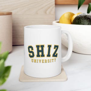 May include: White ceramic mug with a navy blue and gold design. The mug features the text "SHIZ UNIVERSITY" in a collegiate font.