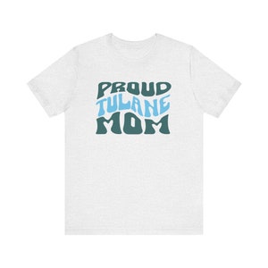 May include: White t-shirt with a blue and green graphic that says "Proud Tulane Mom" in a retro font.