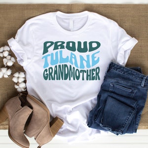 May include: White t-shirt with the text "Proud Tulane Grandmother" in green and blue letters. The shirt is folded and laid on a wooden surface with a pair of brown ankle boots and blue jeans.
