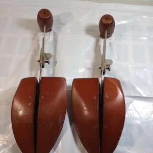 May include: Two wooden shoe trees with metal adjustable arms. The shoe trees are brown and have a rounded shape. The metal arms are silver and have a spring-loaded mechanism.