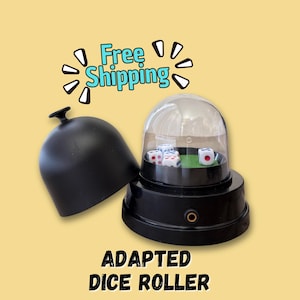Könnte beinhalten: Schwarzer angepasster Würfelroller mit einer transparenten Kuppel, die Würfel auf einer grünen Oberfläche zeigt. Der Würfelroller hat einen schwarzen Deckel und der Text "ADAPTED DICE ROLLER" ist darunter aufgedruckt.