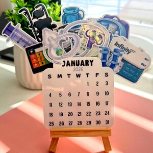 May include: A January 2026 calendar on a small wooden easel, with medical-themed stickers in the background. The stickers include a syringe, medical equipment, and a calculator. The calendar is white with black text.