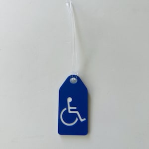 May include: Blue luggage tag with a white wheelchair accessibility symbol. The tag has a clear plastic loop for attaching to luggage.