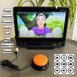 May include: A black tablet computer with a screen showing a video of a woman teaching preschoolers. The tablet is connected to a large orange button with a black base. The button is connected to a black cord. The screen shows the text "Dinosaur eggs."  The tablet has a white label with the text "tobli dynavox" on the top right corner. The screen also shows the text "Preschool & Toddler Learning Video with Ms Rachel Learn Shapes, Letters, Numbers, Colors & Mo". Below the tablet are nine white circles with black icons representing common media player controls.