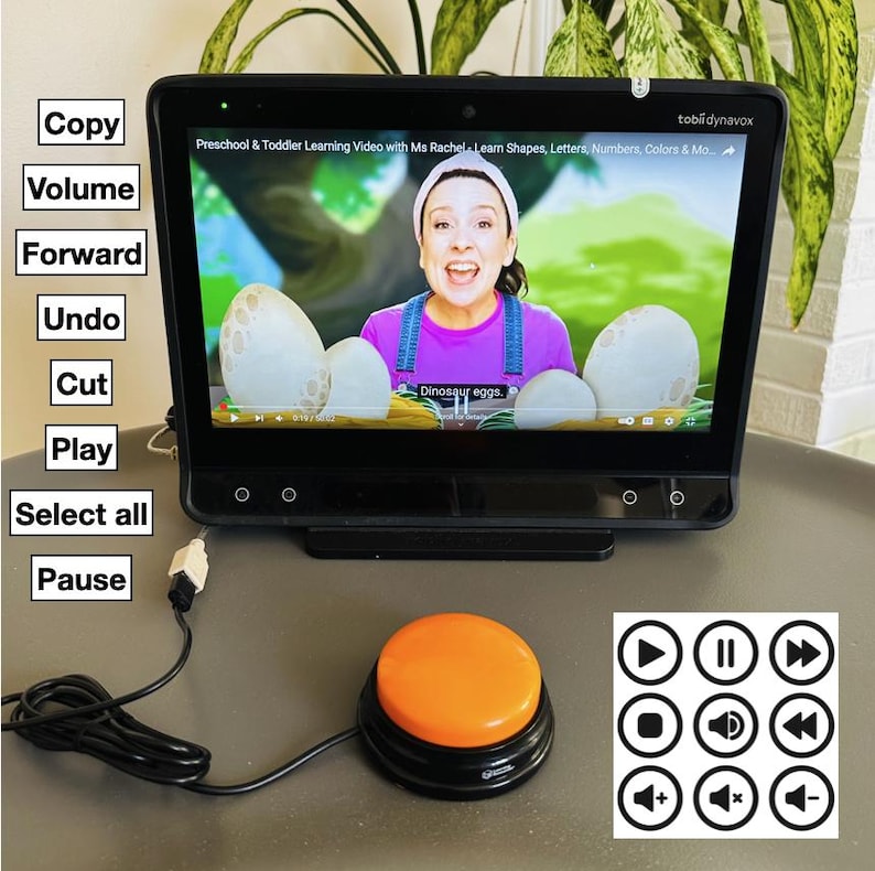 May include: A black tablet computer with a screen showing a video of a woman teaching preschoolers. The tablet is connected to a large orange button with a black base. The button is connected to a black cord. The screen shows the text "Dinosaur eggs."  The tablet has a white label with the text "tobli dynavox" on the top right corner. The screen also shows the text "Preschool & Toddler Learning Video with Ms Rachel Learn Shapes, Letters, Numbers, Colors & Mo". Below the tablet are nine white circles with black icons representing common media player controls.