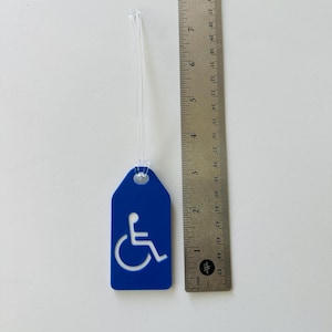 May include: Blue luggage tag with a white wheelchair accessibility symbol. The tag is attached to a clear plastic string.