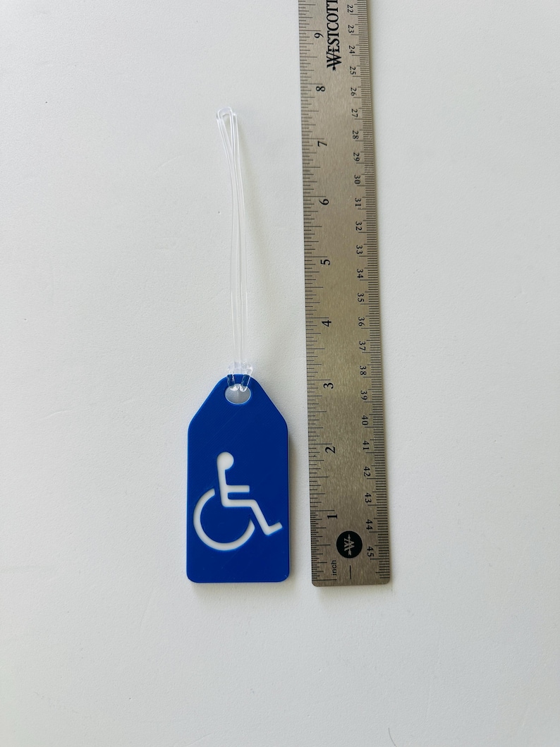 May include: Blue luggage tag with a white wheelchair accessibility symbol. The tag is attached to a clear plastic string.