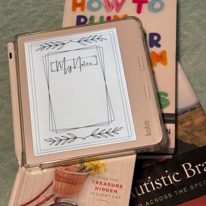 以下が含まれることがあります： ピンクの電子書籍リーダーで、白黒の画面に「My Notes」というテキストが表示されたメモテンプレートが表示されています。電子書籍リーダーは、「Find the treasure hidden in every day」というテキストが書かれたピンクの本と、「Autistic Brains」と「Thinking Across the Spectrum」というテキストが書かれた黒い本のの上に置かれています。