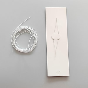Op de afbeelding: Een witte rechthoekige sieraden displaykaart met een uitgesneden ontwerp en een kleine, opgerolde bundel witte draad. De kaart heeft een cirkelvormig gat aan de onderkant. De draad is voor het bevestigen van sieraden.